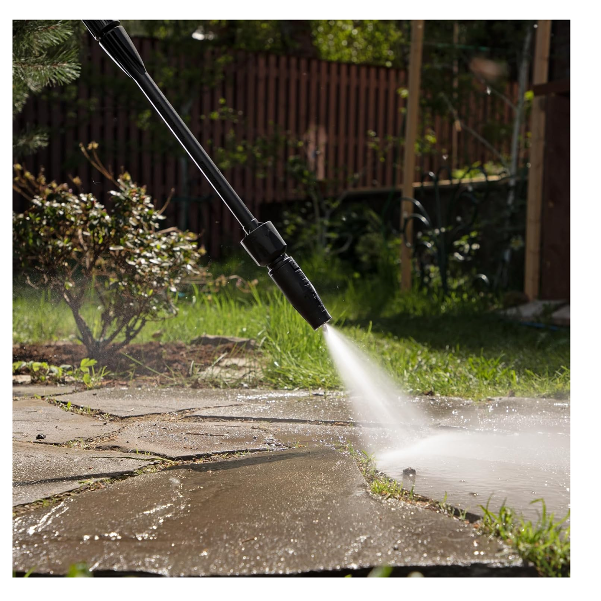 Pressure washer nozzle spraying water onto a stone patio in a garden setting.