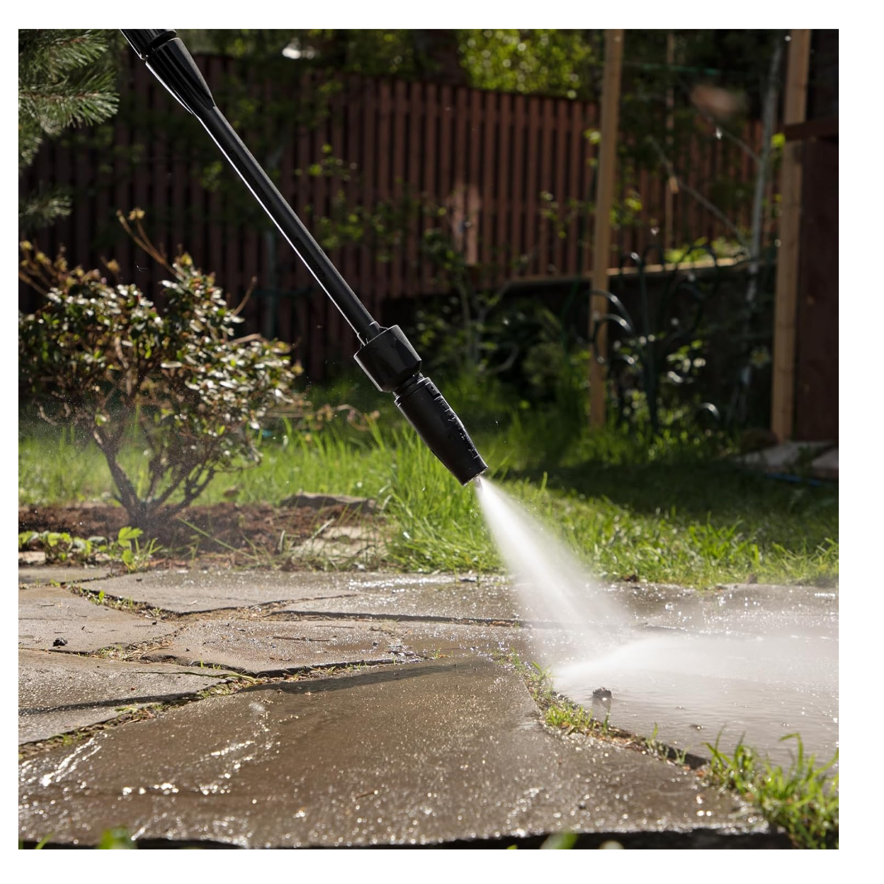 Pressure washer nozzle spraying water onto a stone patio in a garden setting.