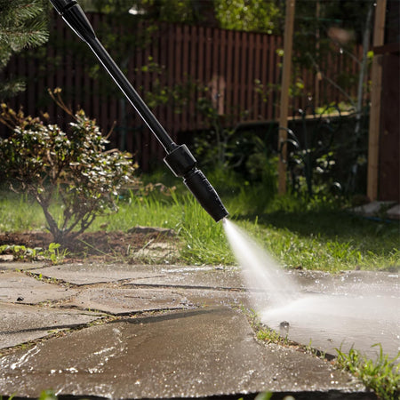 High-pressure washer nozzle spraying water on a stone patio with grass and plants in the background.