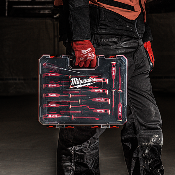 A Milwaukee screwdriver bit set in a clear plastic case with red tools, held by a person wearing black work pants and red Milwaukee gloves in a dark workshop setting.
