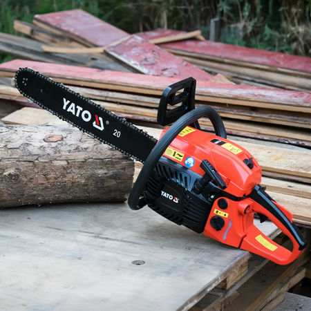 A YATO 20-inch chainsaw with a bright orange body and black bar, resting on a wooden pallet with stacked lumber in the background.